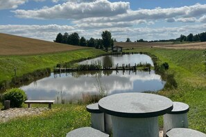 Un cadre extérieur paisible avec un étang, un banc en bois et une table en pierre entourée de chaises.