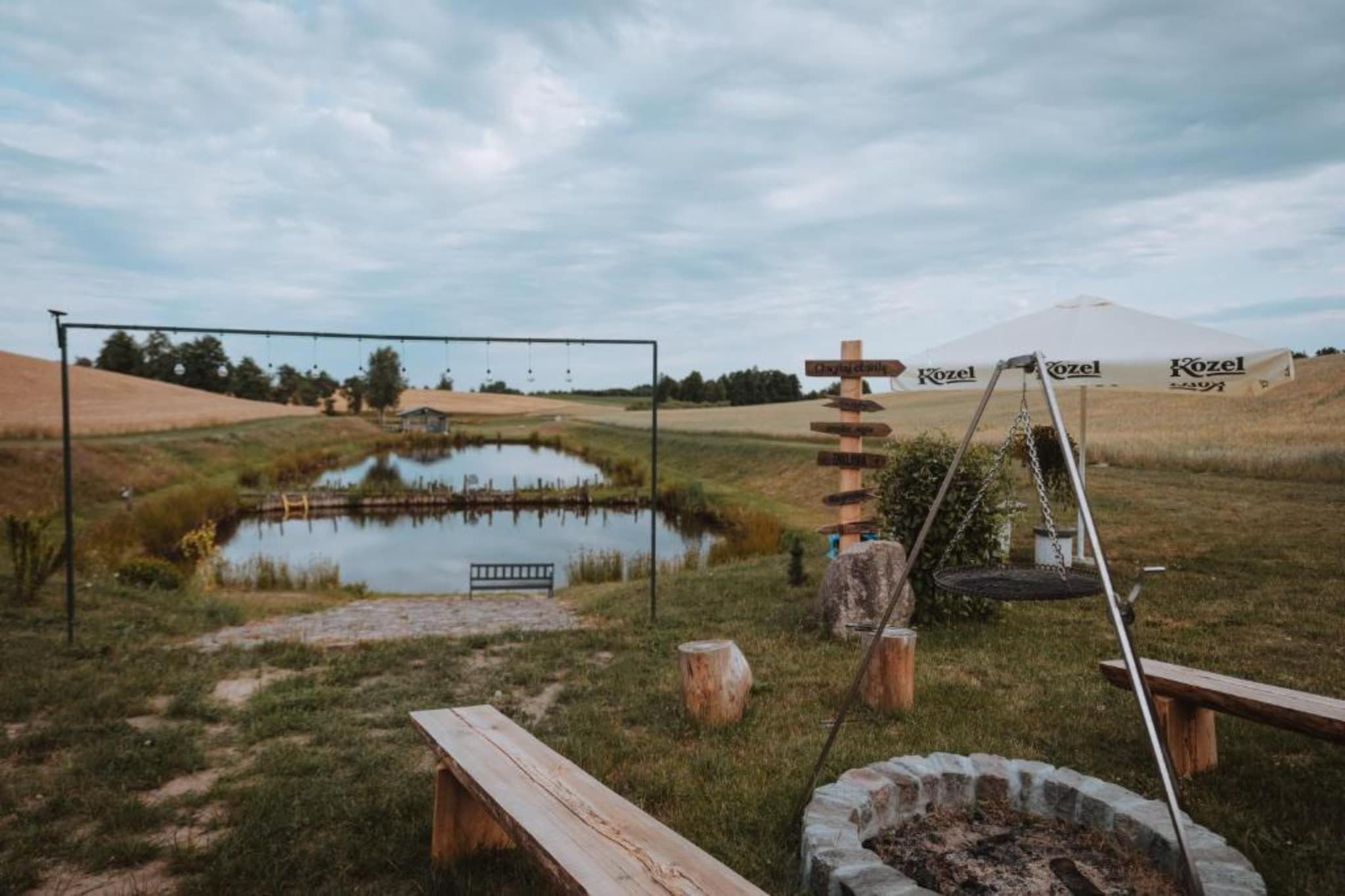 A rustic outdoor setting with a pond, wooden benches, a swing set, and a Kozel tent in the background.