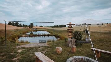 A rustic outdoor setting with a pond, wooden benches, a swing set, and a Kozel tent in the background.