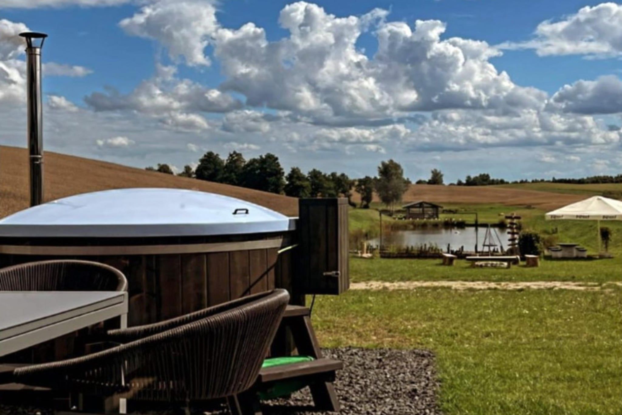 Een hottub met een houten terras en stoelen, een houten omheining, en een vijver met een schommel in een grasveld.