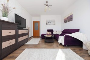 Moderni olohuone, jossa on televisio, tumma puinen kaappi, violetti sohva ja pieni pyöreä pöytä, jonka päällä on koriste-esineitä.