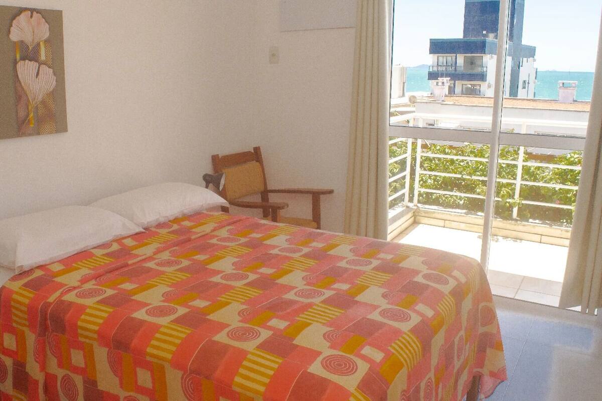Un dormitorio con cama, una silla, una mesita de noche, un cuadro y un balcón con vistas al mar y edificios.