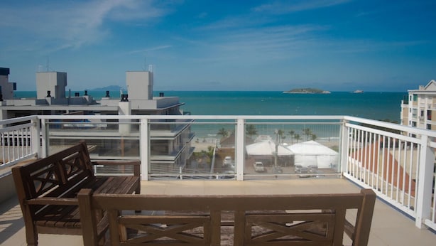 A balcony with a wooden table and bench, overlooking a beach with umbrellas and a clear blue sky. - Mirante dos Ingleses (Florianopolis)