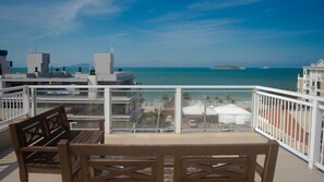 Un balcón con una mesa y banco de madera, con vista a una playa con sombrillas y un cielo azul despejado.