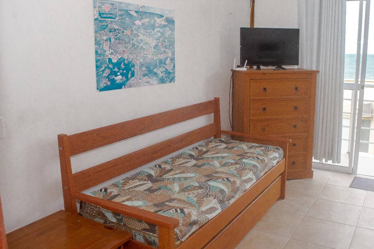 Un dormitorio con una cama de madera, un tocador, un televisor y vista al mar a través de un ventanal corredera.