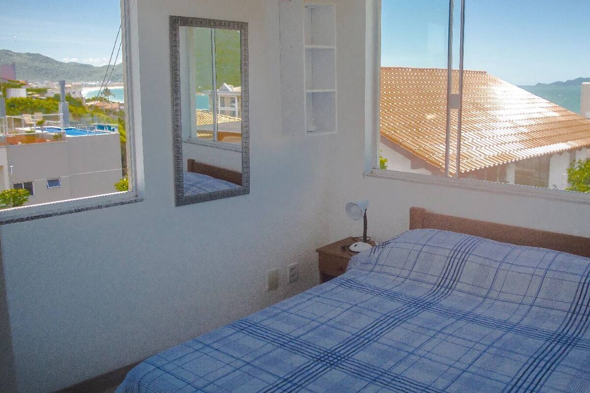 Un dormitorio con cama, mesita de noche, espejo y ventana con vistas a edificios y la playa.