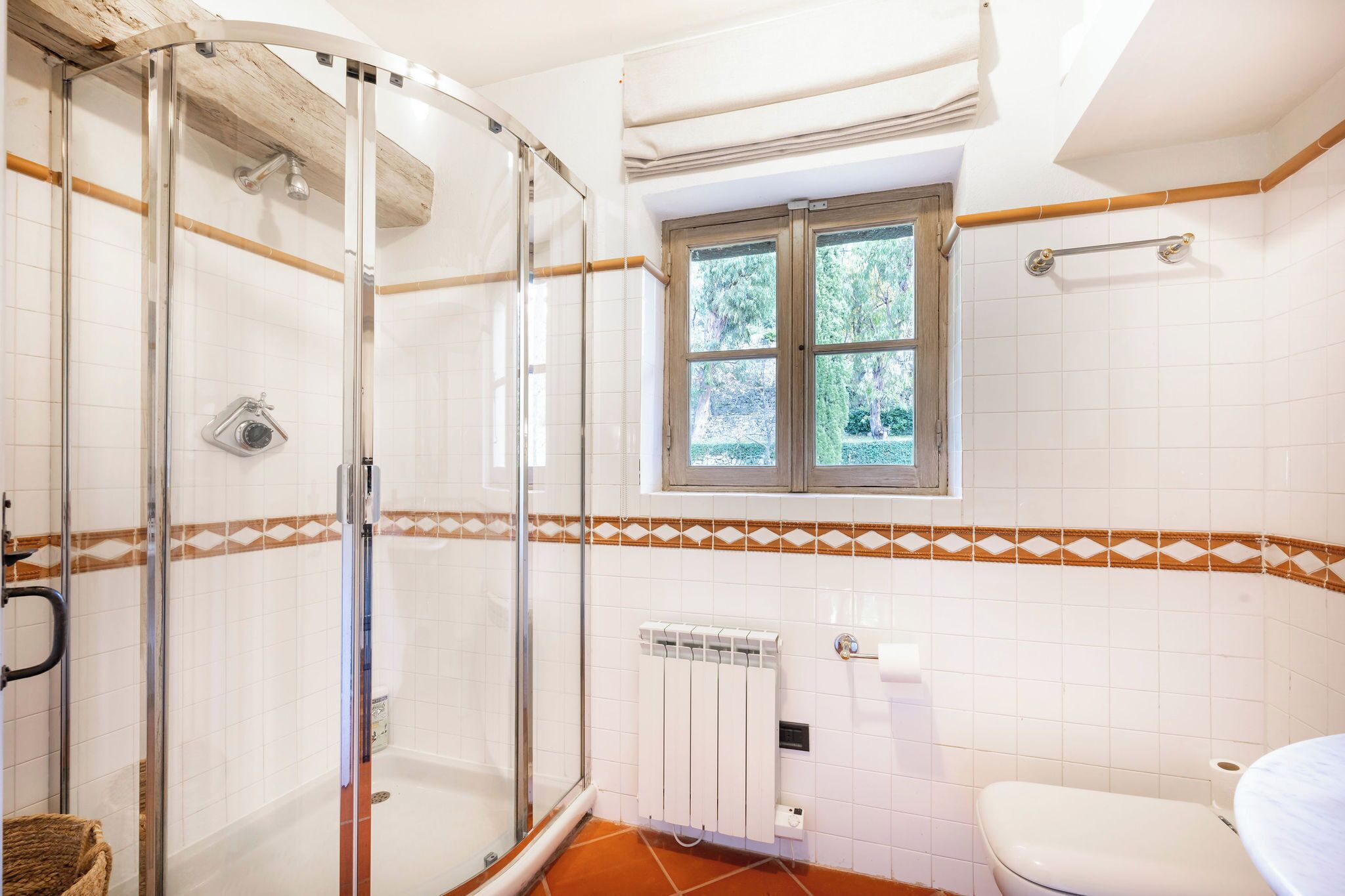 A bathroom with a glass shower enclosure, a toilet, a window with a view of greenery, and a radiator.