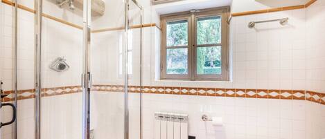 A bathroom with a glass shower enclosure, a toilet, a window with a view of greenery, and a radiator.