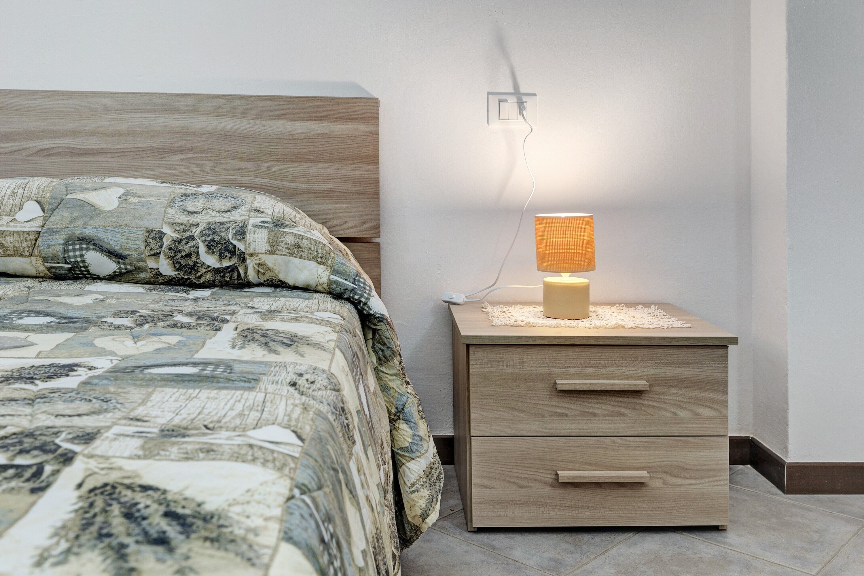 A neatly made bed with a patterned bedspread, a bedside table with a lamp, and a wall socket.
