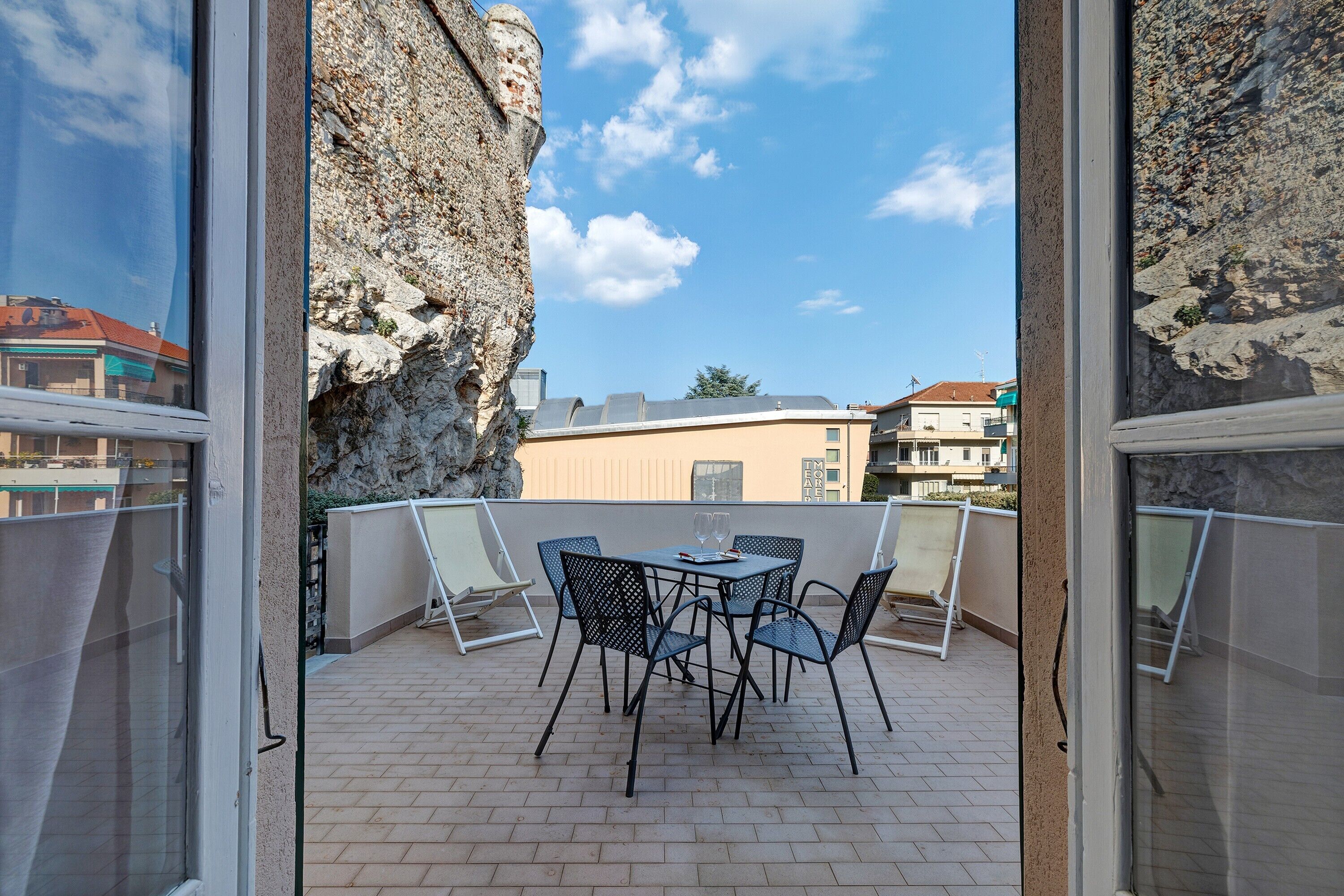 A balcony with a table and chairs, a view of buildings, and a rocky cliff.