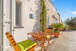 A wooden table and chairs outside a building with a white door and shutters, potted plants, and a cobblestone path. - Apartment 'Casa Di Gaia' with Wi-Fi and Air Conditioning (Carloforte)