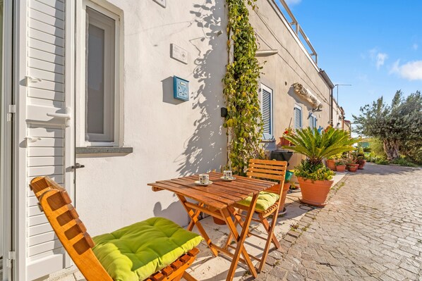 A wooden table and chairs outside a building with a white door and shutters, potted plants, and a cobblestone path. - Apartment 'Casa Di Gaia' with Wi-Fi and Air Conditioning (Carloforte)