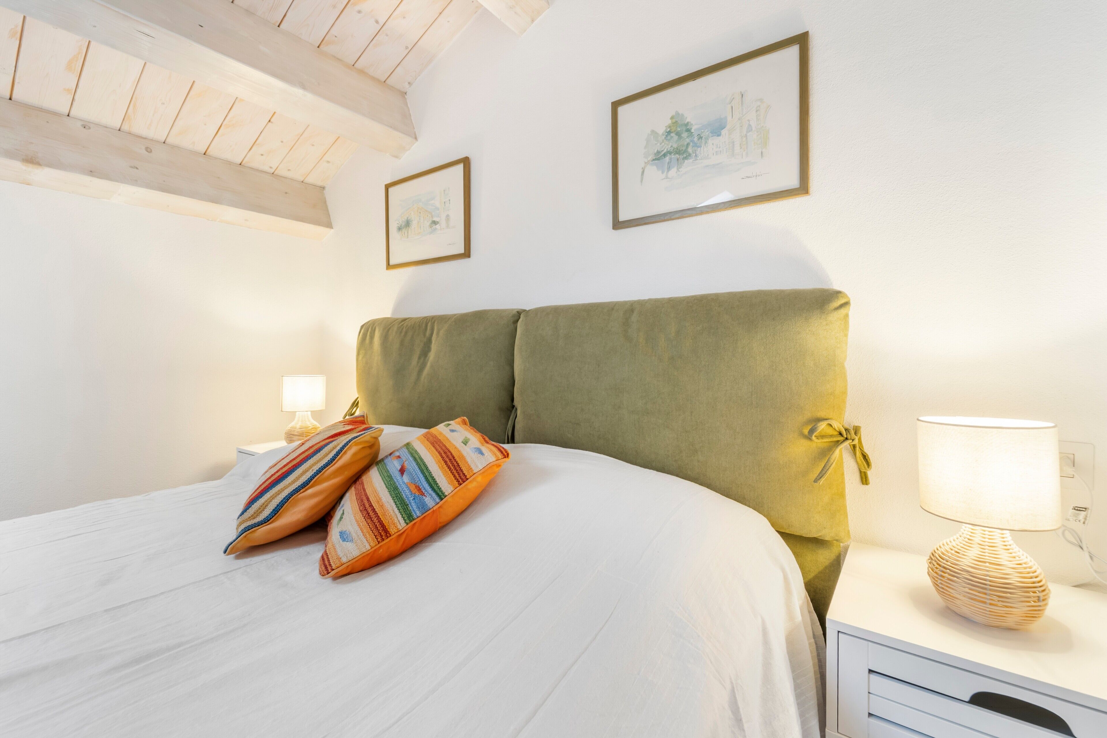Una habitación con una cama, dos cuadros en la pared, una lámpara en una mesita de noche y una ventana con vigas de madera.