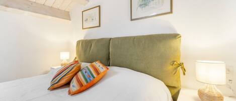 Una habitación con una cama, dos cuadros en la pared, una lámpara en una mesita de noche y una ventana con vigas de madera.