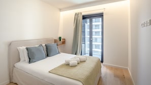 Ein Schlafzimmer mit Bett, Kissen, Decke, Nachttisch, Fenster und Blick auf Gebäude.