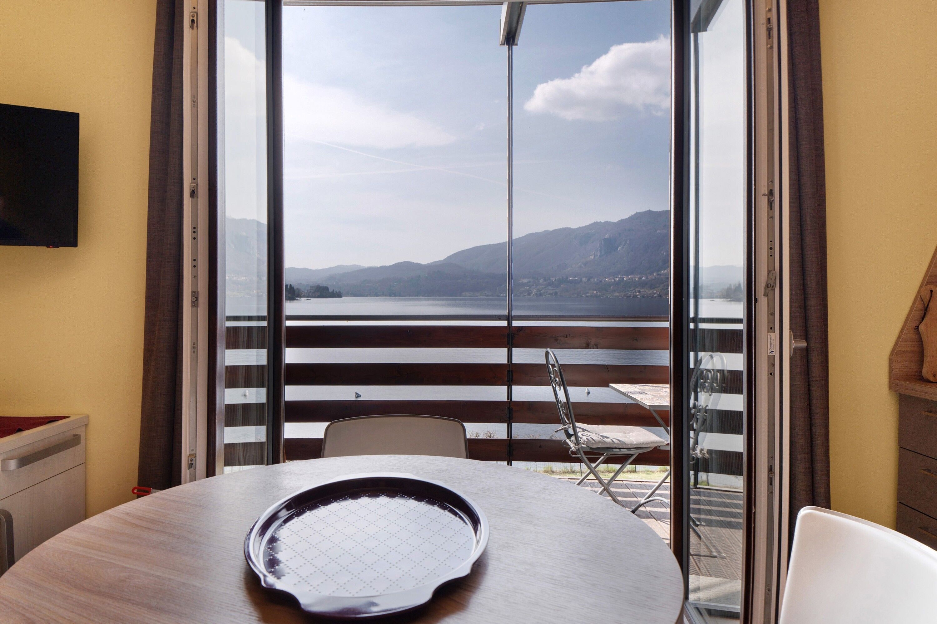 A room with a table, chairs, a TV, and a view of a lake and mountains through a sliding door.