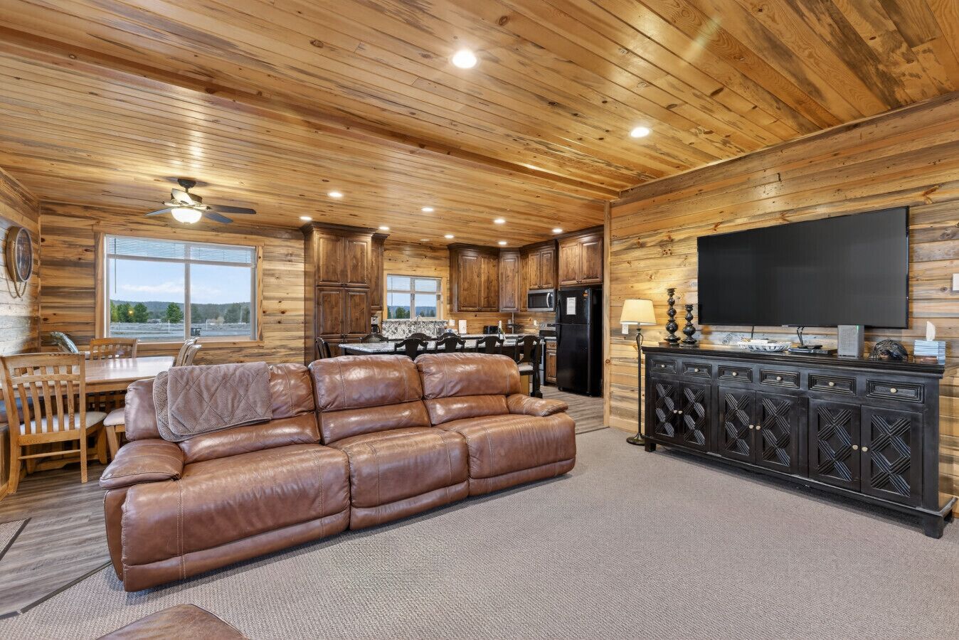 Ein Interieur einer Holzblockhütte mit Ledersofa, Esstisch, Küchenbereich und einem Flachbildfernseher.