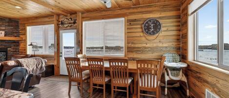 A cabin interior with a wooden ceiling, a dining table with chairs, a fireplace, and a clock on the wall.