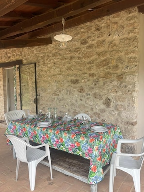 Un intérieur avec des murs en pierre, une table dressée pour le repas, des chaises en plastique blanc et un luminaire suspendu.