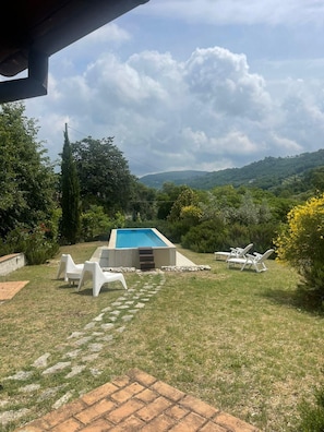 Une piscine entourée de chaises longues, bordée d’une pelouse et d’arbres, avec un paysage vallonné en arrière-plan.