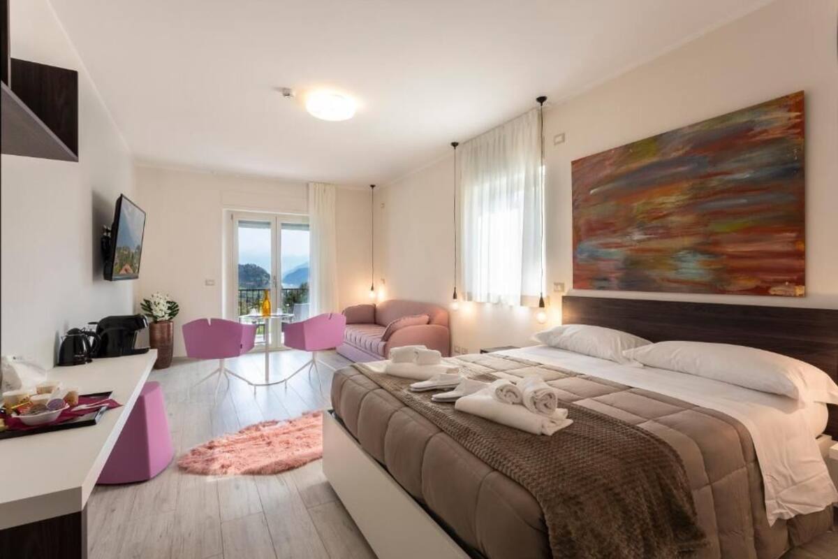 A modern hotel room with a bed, a desk, a chair, a sofa, and a large window.