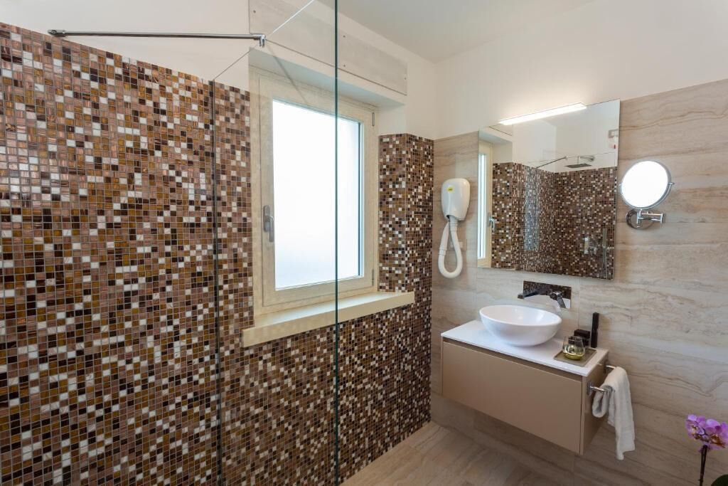 A bathroom with mosaic tile walls, a large window, a sink, a mirror, and a hairdryer.