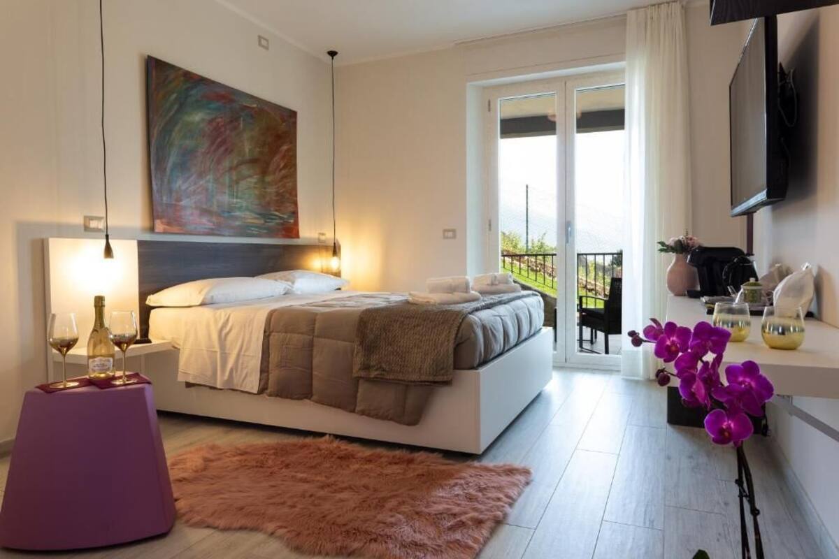 A modern bedroom with a bed, bedside table with glasses, a painting, a balcony with plants, and a TV.