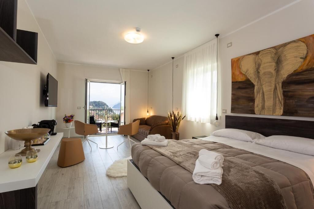 A modern hotel room with a large bed, a desk with a flat-screen TV, a dining area with chairs, and a balcony with a view.