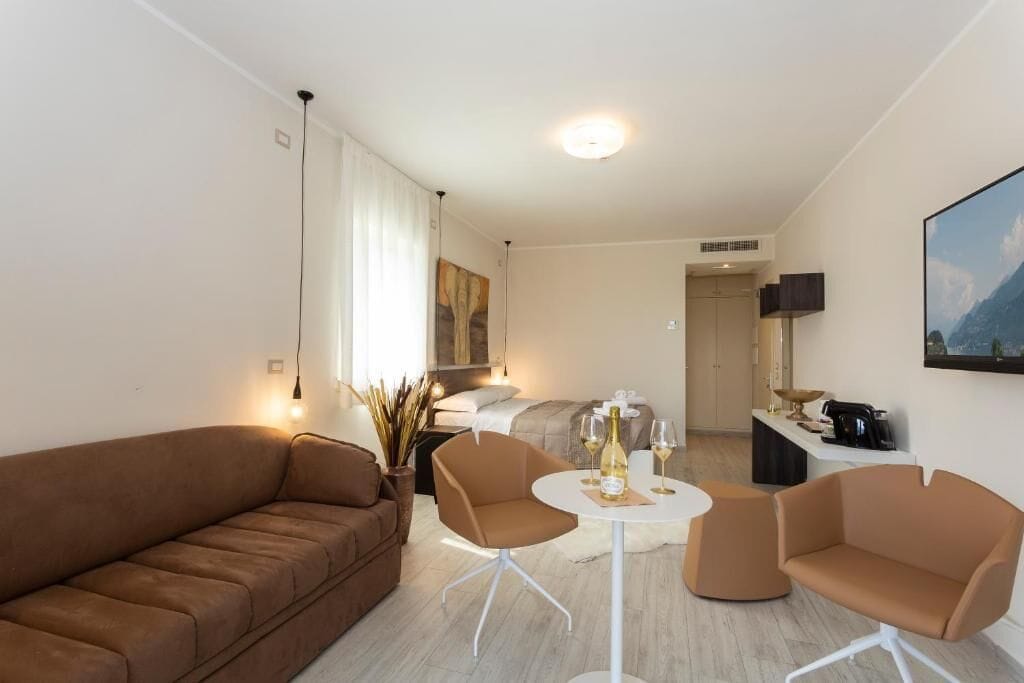 Ein modernes Hotelzimmer mit einem braunen Sofa, zwei Sesseln, einem kleinen runden Tisch mit Flasche und Gläsern, einem Bett mit weißem Kopfteil, einem an der Wand montierten Fernseher und einem Gemälde über dem Bett.
