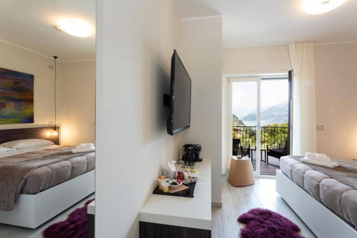 A modern hotel room with two beds, a flat-screen TV, a small table with items, and a balcony with a view.
