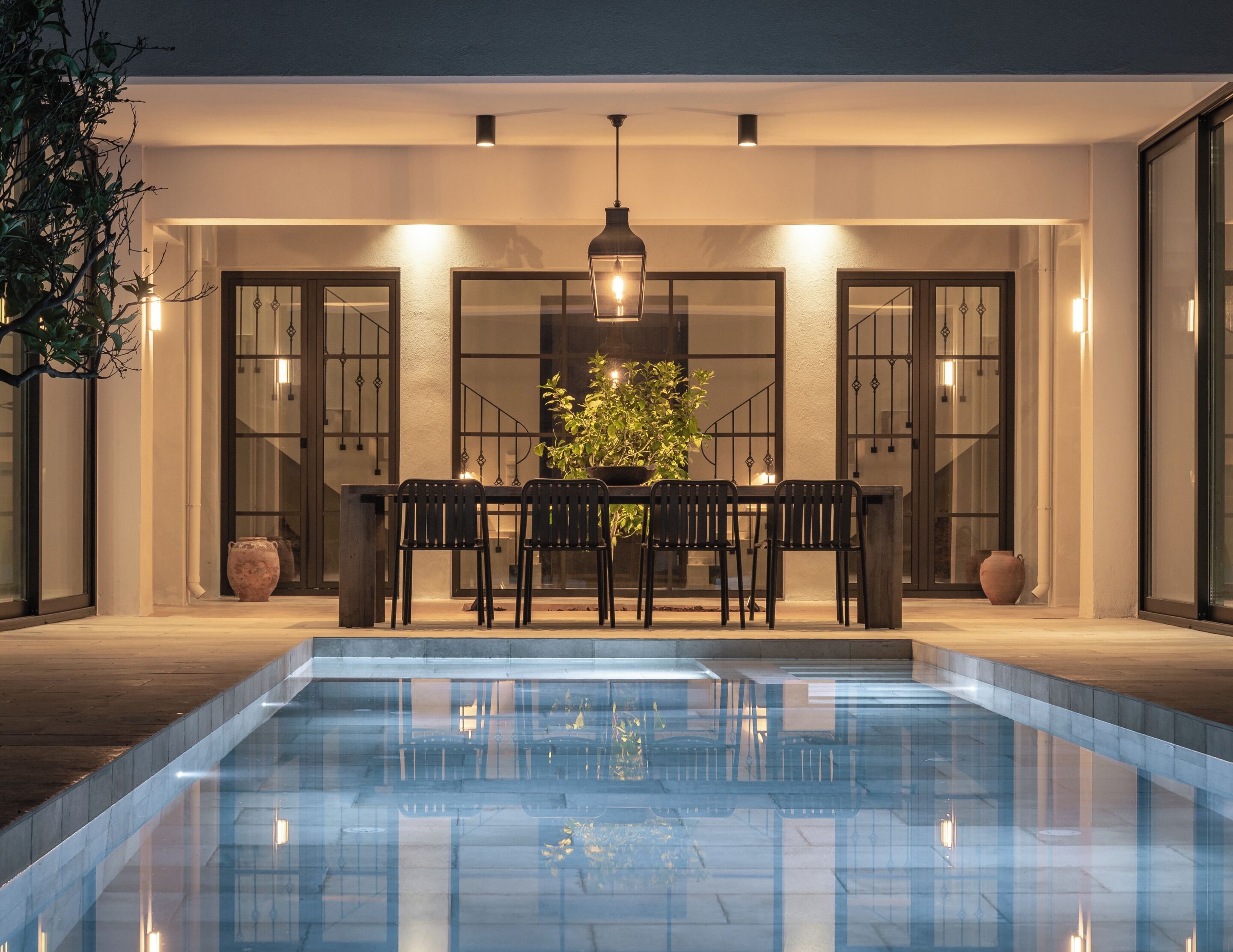 A poolside dining area with a table and chairs, a hanging light fixture, and potted plants.