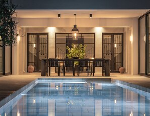 A poolside dining area with a table and chairs, a hanging light fixture, and potted plants. - Citrus Grove Luxury Villas (Rethymno)