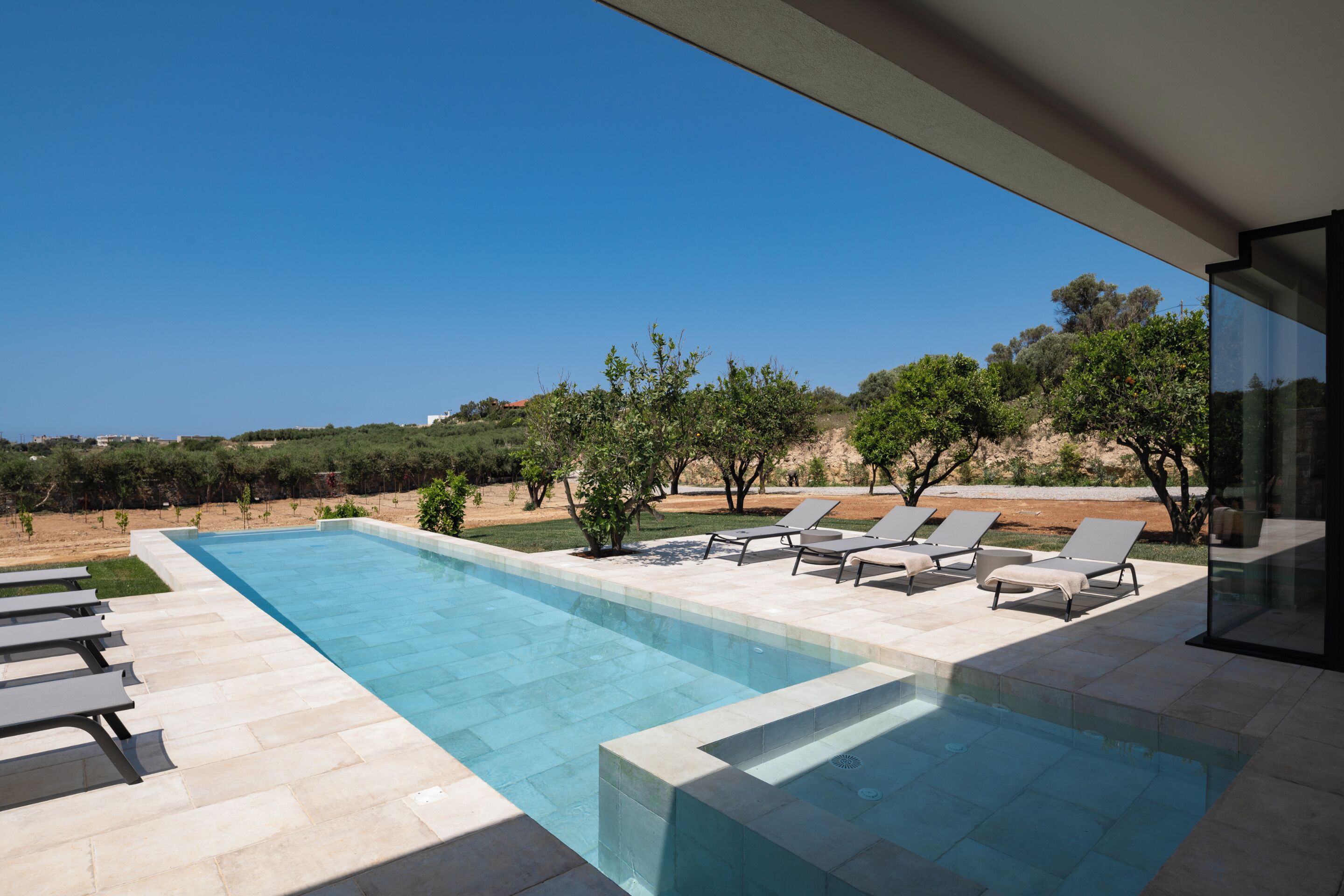 A modern outdoor pool area with clear blue water, surrounded by lounge chairs and a view of the landscape.