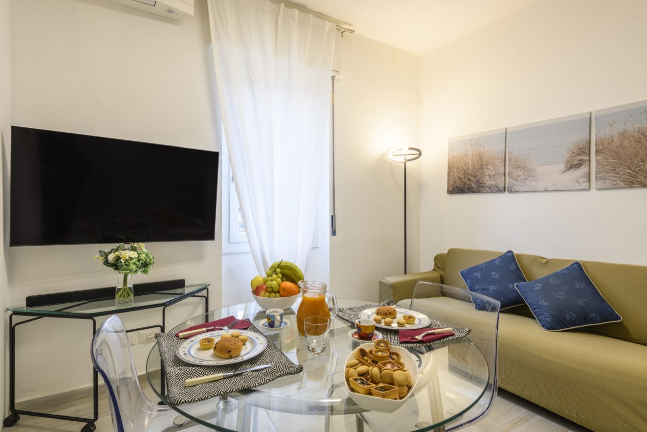 Een woonkamer met een glazen salontafel, een bank, een flatscreen-televisie en een schilderij aan de muur.