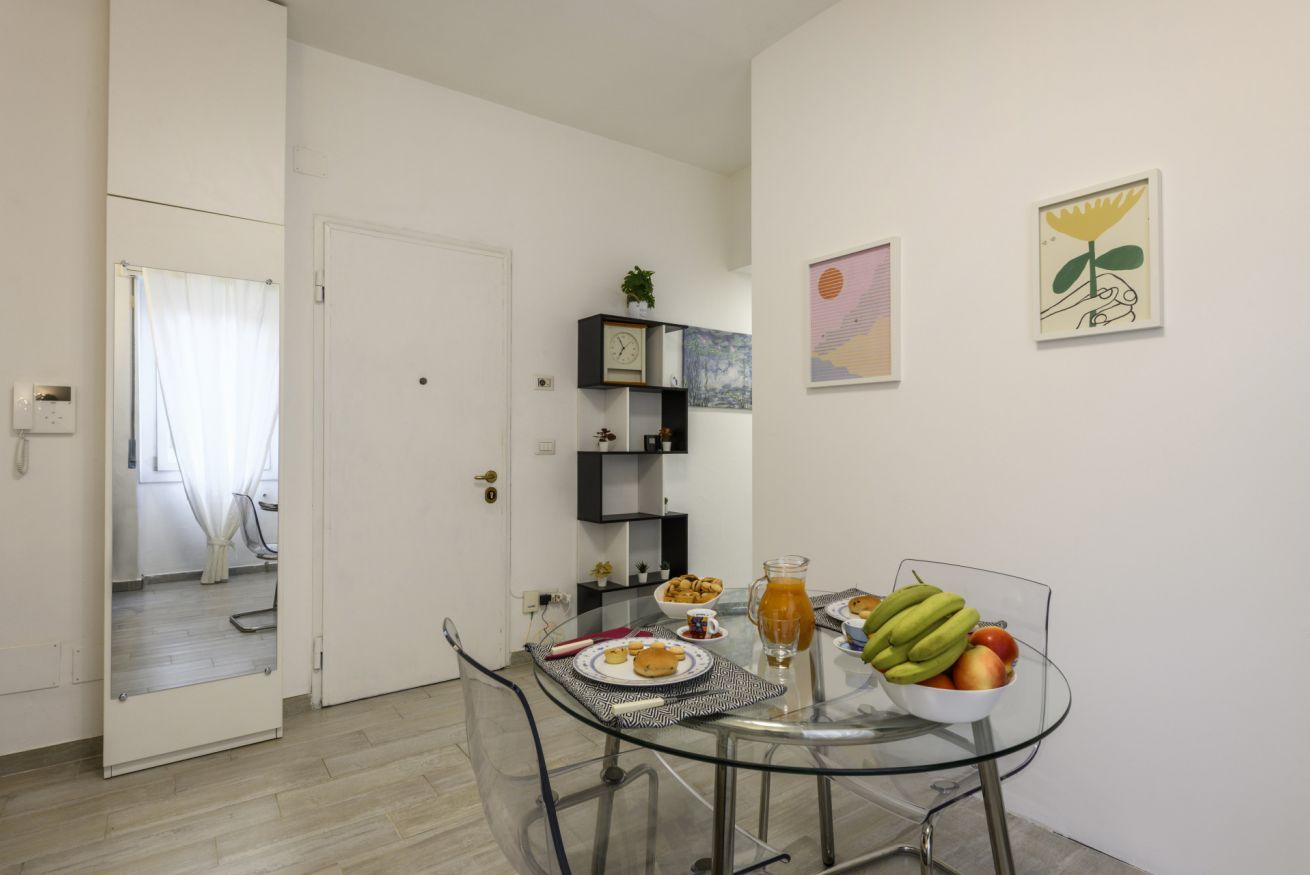 Un espace repas moderne avec une table en verre, une porte blanche et une petite étagère noire agrémentée d’une plante.