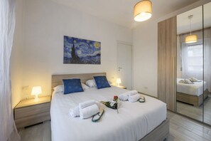 A neatly made bed with towels, a bottle of champagne, and slippers. There is a painting on the wall and a mirror reflecting the room. - Monte Ceneri, 71 — Modern, practical, and well-connected (Milan)
