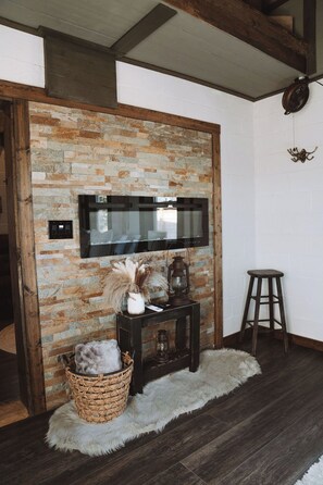 A room with a stone wall, a mounted flat-screen TV, a wicker basket with a fluffy object, a small wooden stool, and a fur rug on the floor. - Luxury Renovated Barn Near Bennett Springs – Cozy & Private (Lebanon)