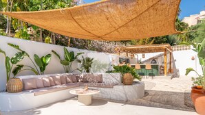 Uma área de estar ao ar livre com sol, cobertura de tecido trançado, um banco acolchoado, uma mesa redonda e plantas em vasos.