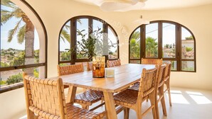 Uma área de refeições com mesa e cadeiras de madeira, grandes janelas em arco e vista para palmeiras e edifícios.