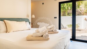 Cama bem arrumada com toalhas dobradas, luminária pendente e vista para o exterior através de uma porta de vidro.