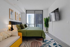 A modern bedroom with a bed, a sofa, a TV, and a large window with a city view. - Downtown Miami 2BR Skyline Bay View Balcony Corner Suite | Luxe Pad (Miami)