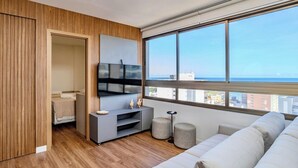 A modern living room with a large window, a flat-screen TV, a grey sofa, and a wooden floor. - 2 suites on a high floor in Barra with ocean view (Salvador)