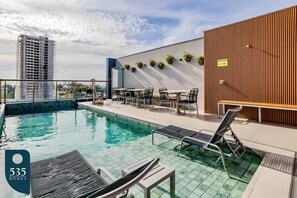 A rooftop pool area with lounge chairs, a wooden wall with potted plants, and a view of a tall building in the background. - 2 suites on a high floor in Barra with ocean view (Salvador)