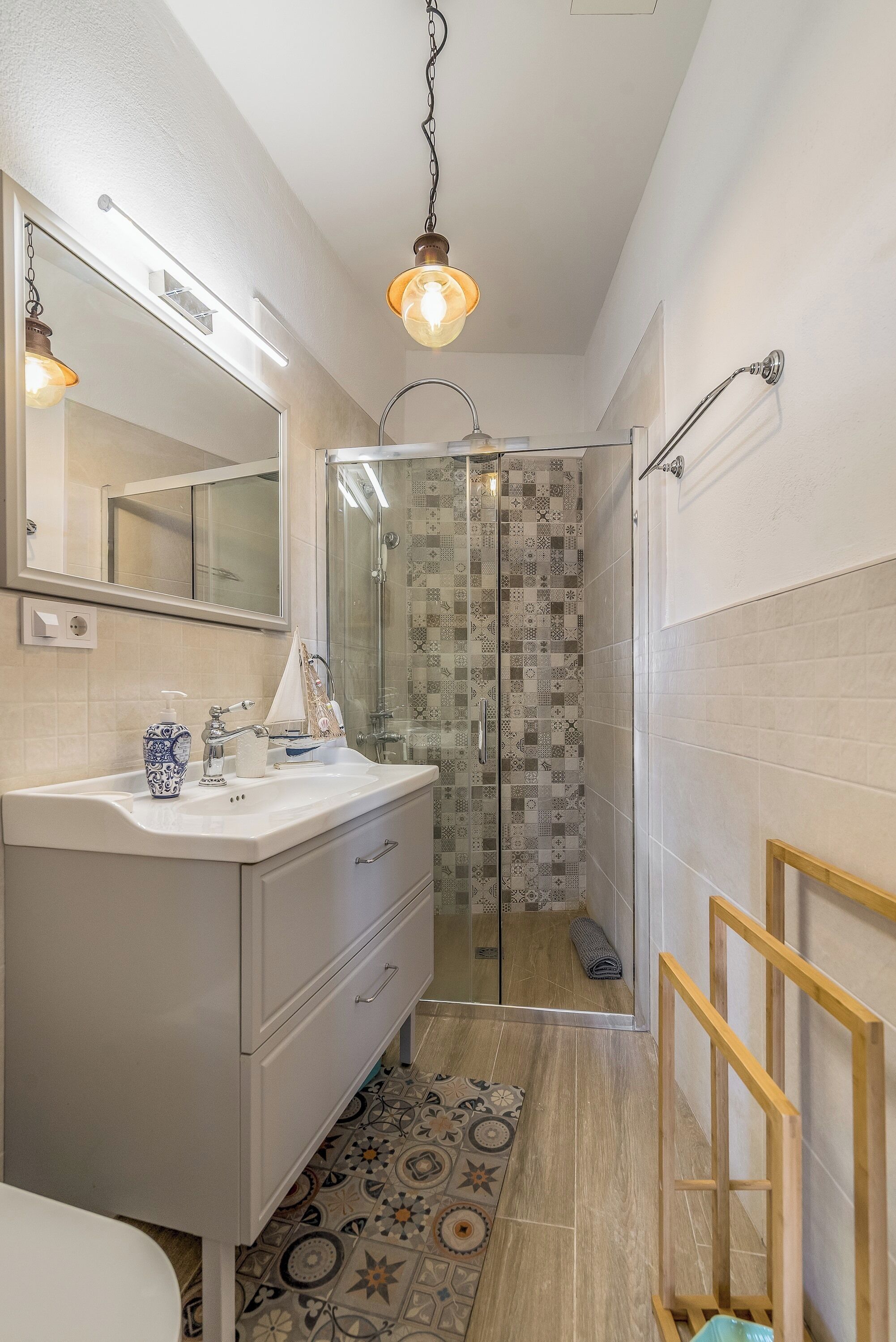 Une salle de bain avec un meuble-lavabo blanc, un miroir, une douche aux carreaux décorés et un luminaire suspendu.