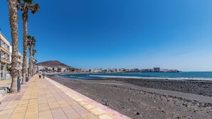 Een kustpad met een strand, palmbomen en gebouwen op de achtergrond.