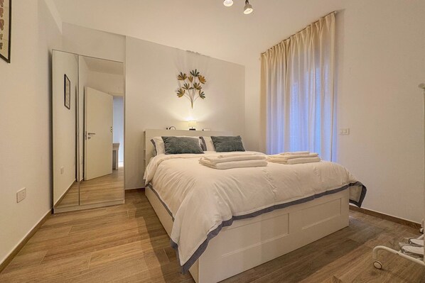 Ein Schlafzimmer mit Bett, Nachttisch, Spiegel, Wandlampe und einem Fenster mit Vorhängen.