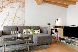 A modern living room with a grey sofa, a glass coffee table, a flat-screen TV, and a bookshelf.