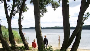 A lakeside view with trees, a stone path, and a distant house. - Mobile Home im Ferienpark mit Seezugang (Dabel)