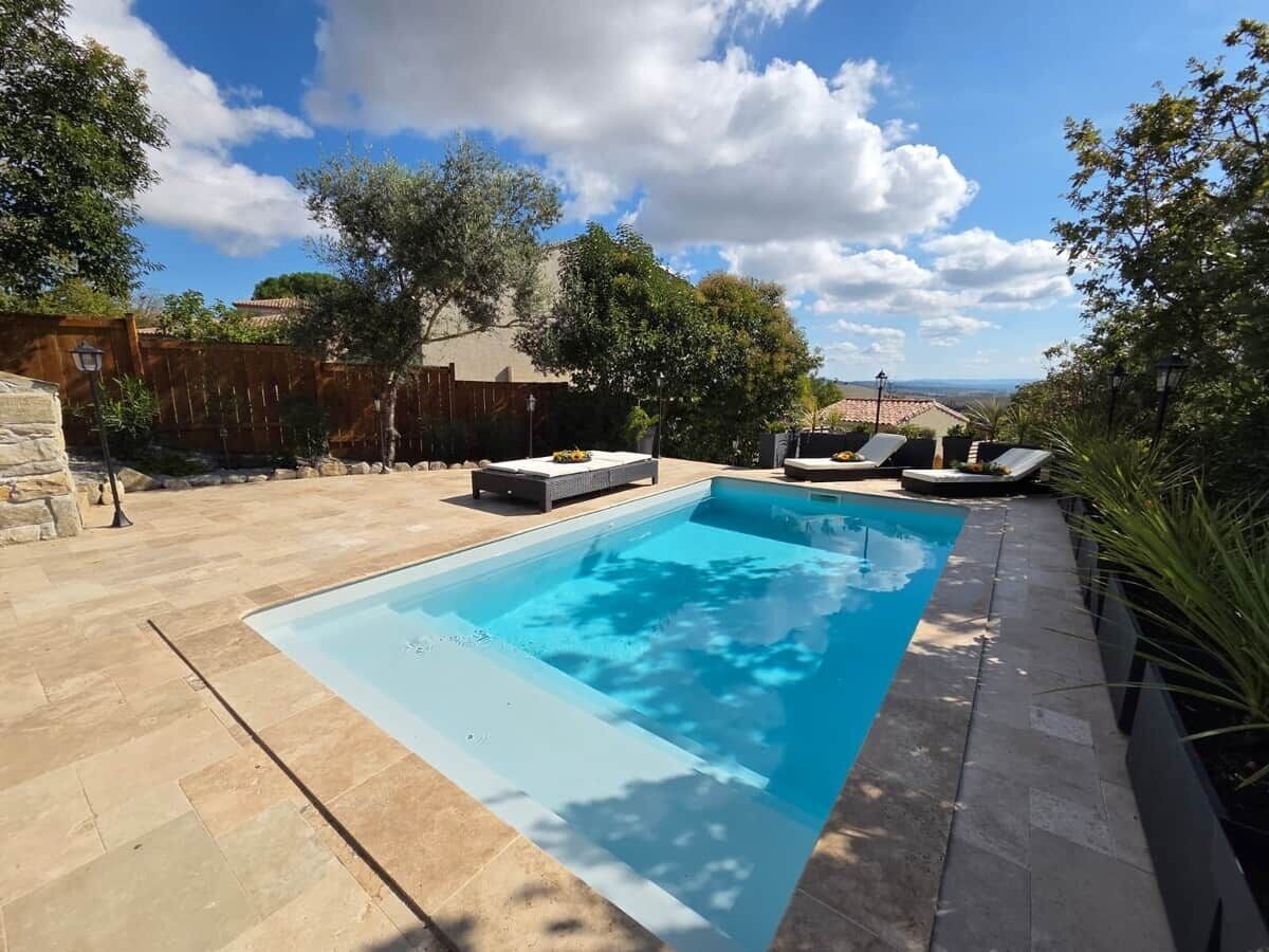 A rectangular swimming pool with a stone deck surrounding it, a wooden fence, and a view of distant hills.