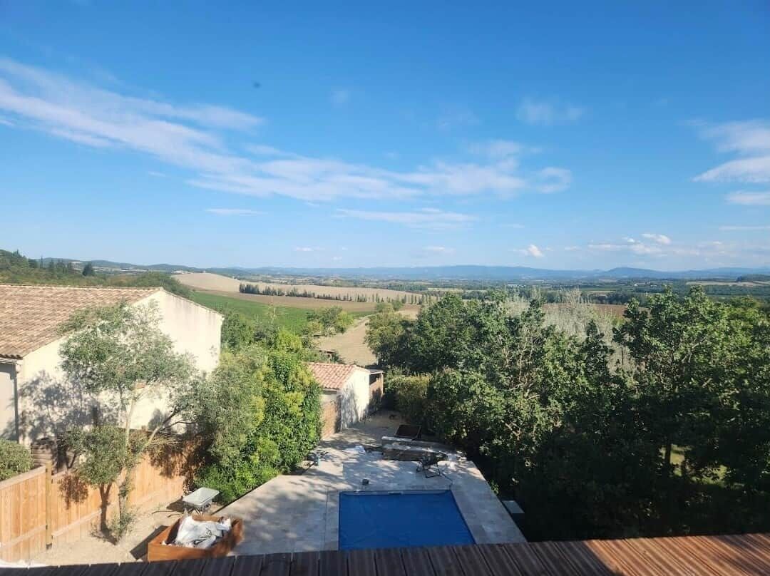 Une piscine dotée d’une terrasse en bois, entourée de verdure et offrant une vue sur des champs lointains.